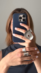 A woman taking a mirror selfie in a room, wearing a dark navy sleeveless top and holding a dark blue phone with a silver ring stand. The background shows a plain light gray wall and a large curved mirror.