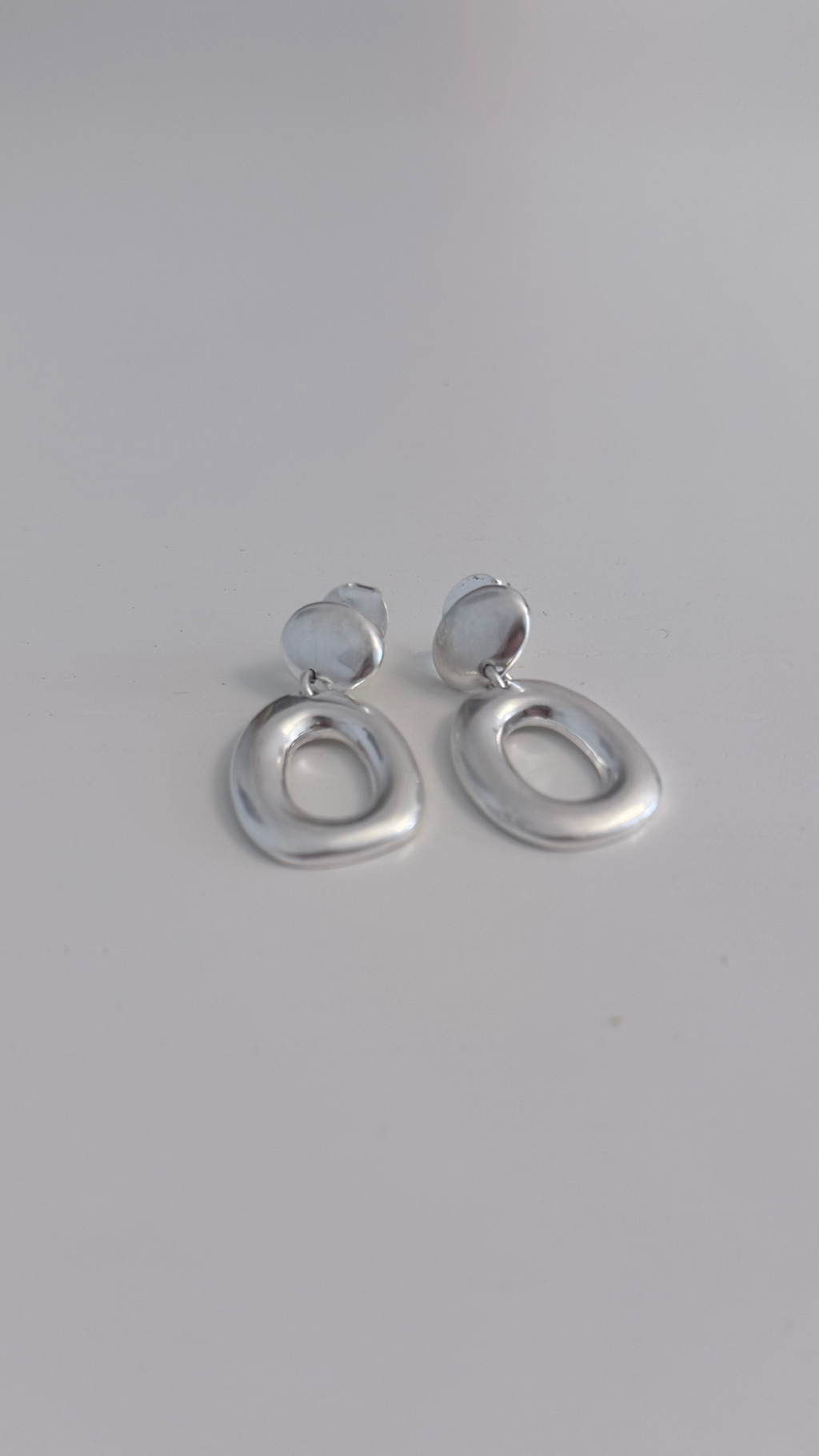 A pair of silver-toned abstract oval drop earrings with a small circular top loop, resting on a plain light gray surface.