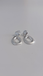 A pair of silver-toned abstract oval drop earrings with a small circular top loop, resting on a plain light gray surface.