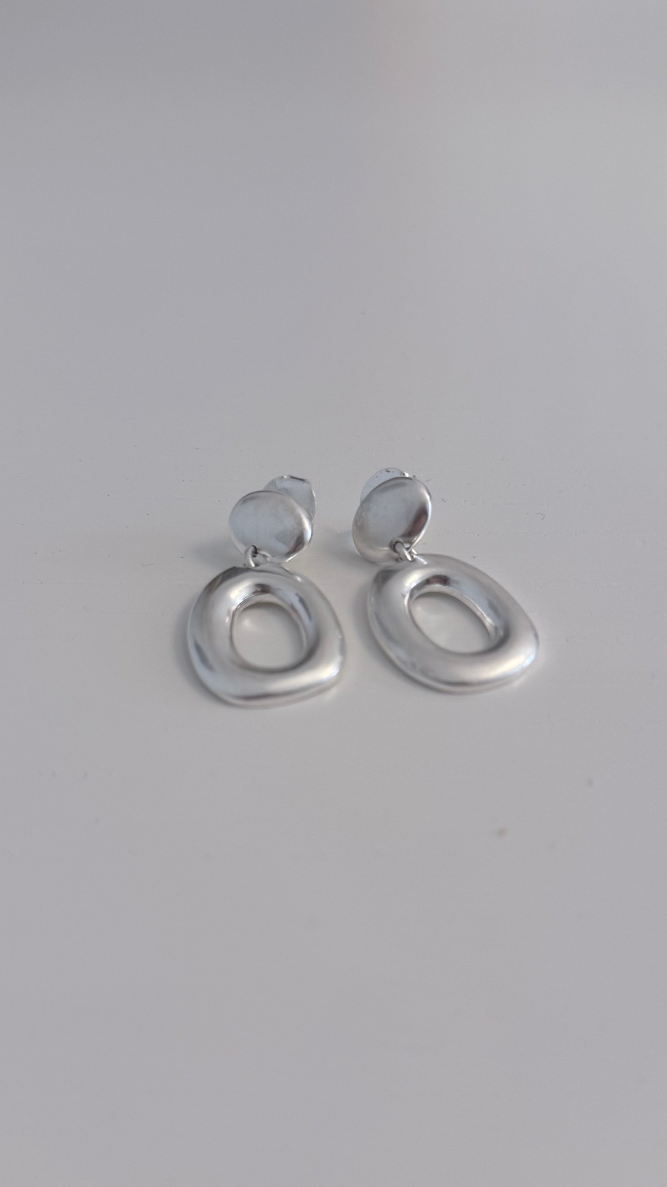 A pair of silver-toned abstract oval drop earrings with a small circular top loop, resting on a plain light gray surface.
