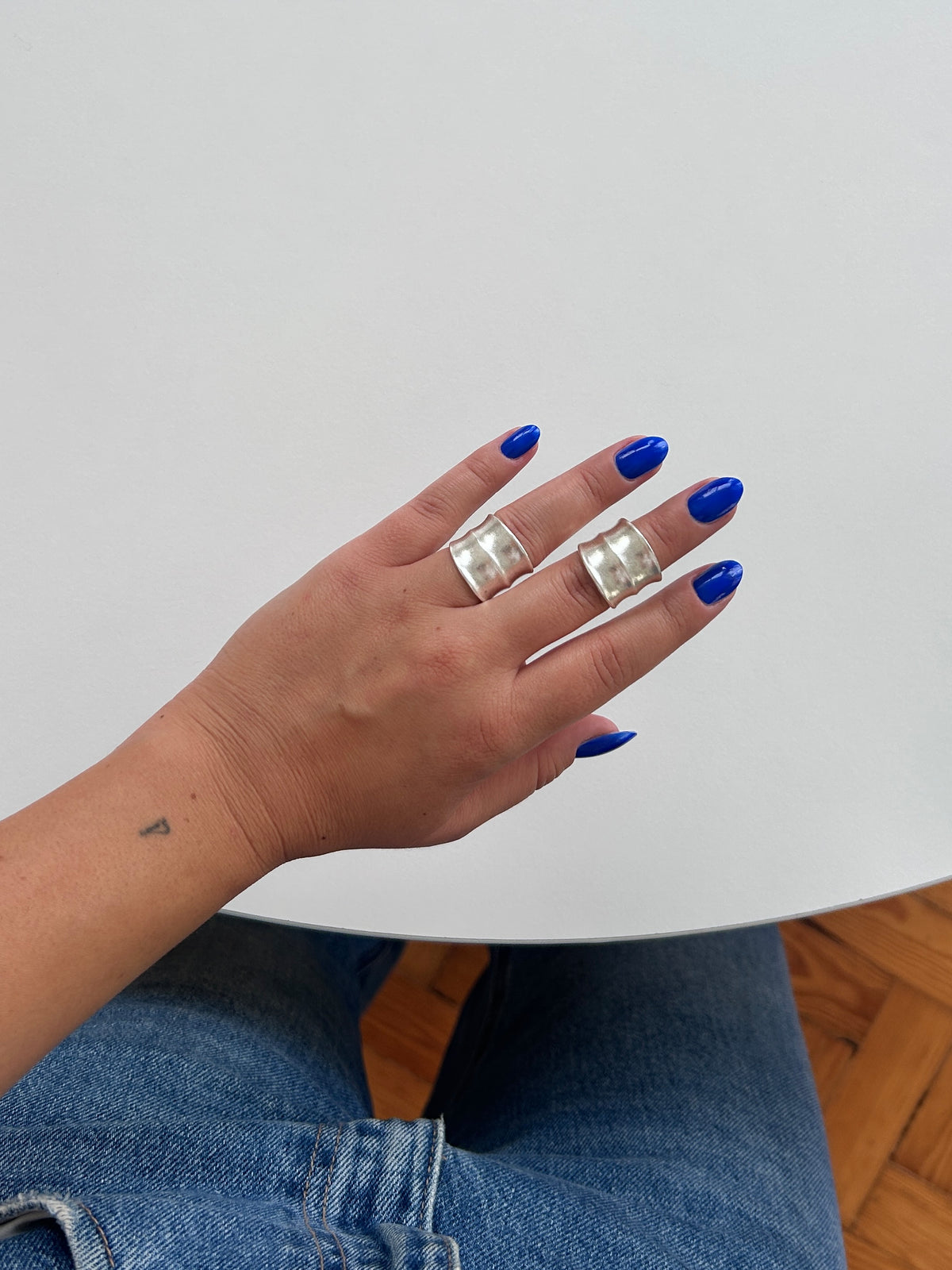 A close-up of a person's hand with bright blue nail polish wearing two chunky silver rings, with blue jeans and a white table visible in the background.
