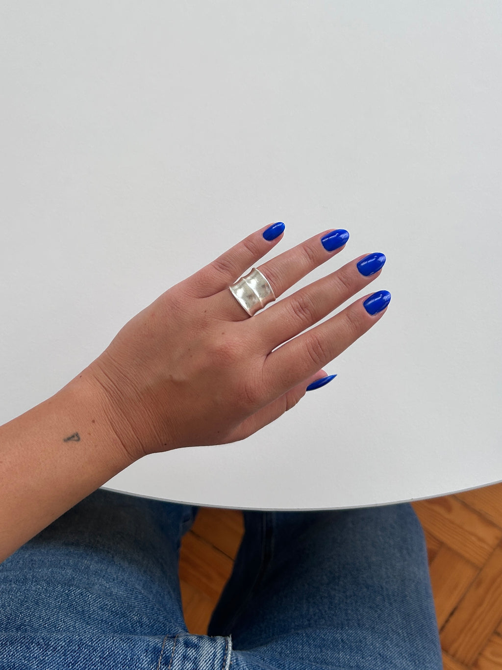 A close-up of a person's hand with bright blue nail polish wearing a wide, chunky silver ring, set against a minimalist white background with a hint of blue jeans and wooden flooring.
