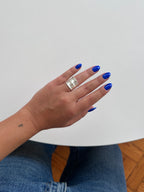 A close-up of a person's hand with bright blue nail polish wearing a wide, chunky silver ring, set against a minimalist white background with a hint of blue jeans and wooden flooring.