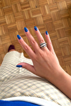A close-up of a hand with blue nail polish wearing a wide silver ring with black zebra-like stripes, resting on a beige plaid blanket over a wooden parquet floor.