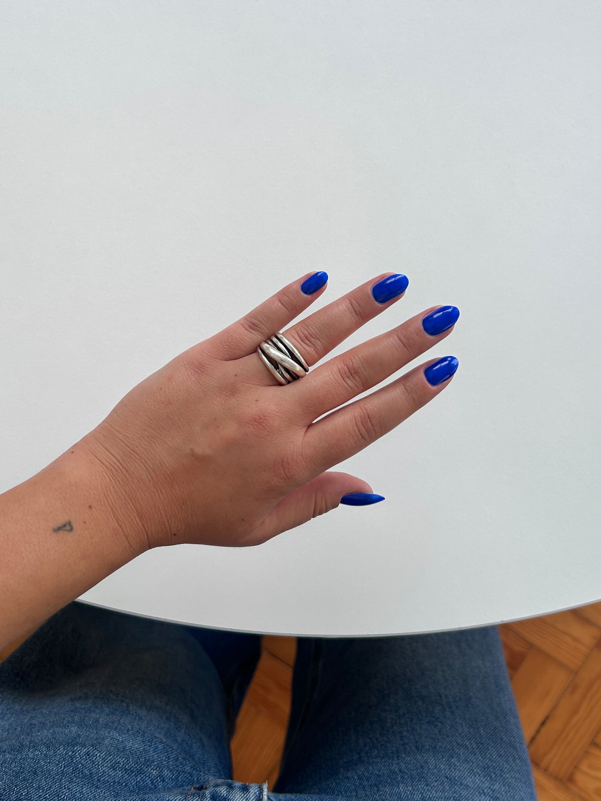 A close-up of a person's hand with bright blue nail polish wearing a wide silver ring with black diagonal stripes, resting on a white table.