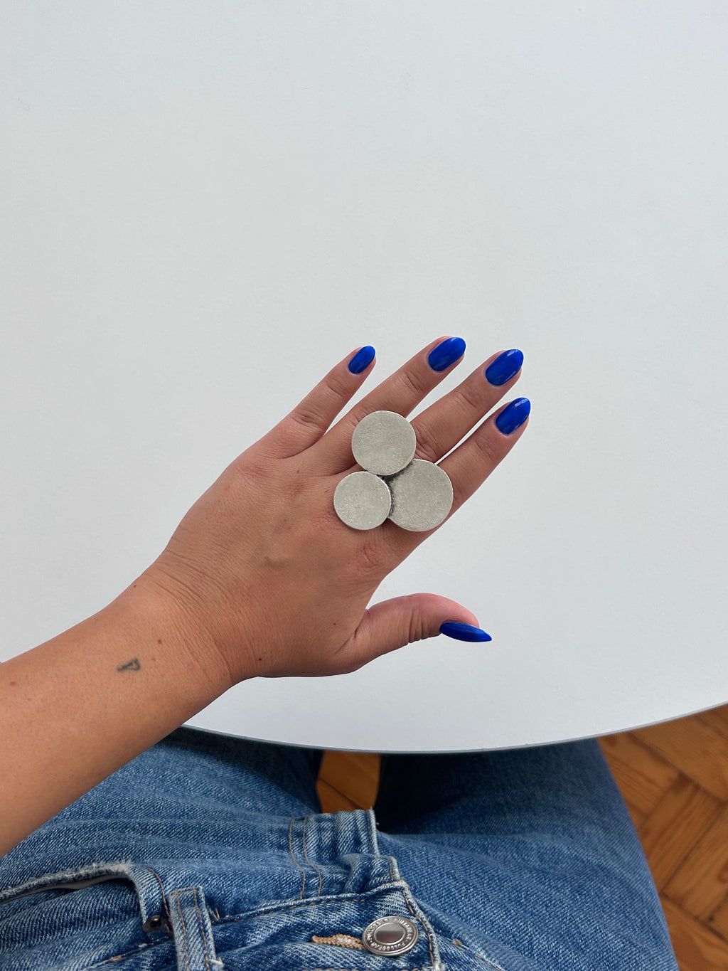 A hand with bright blue nail polish holds three round, silver-toned metal discs against a minimalist white background.